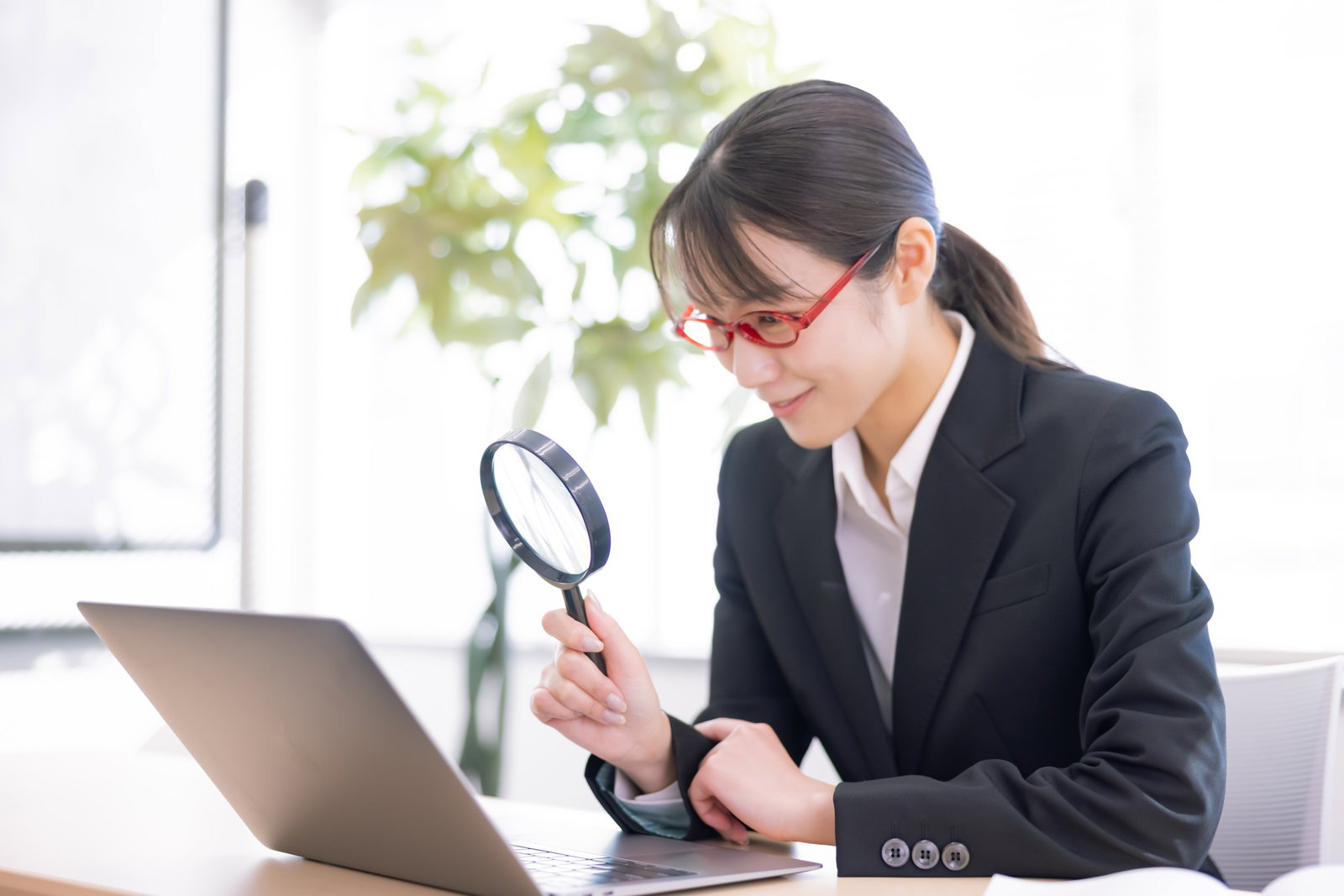 虫眼鏡を手に持ち、覗き込みながらほくそ笑む表情の女性会社員［モデル：SAKI］