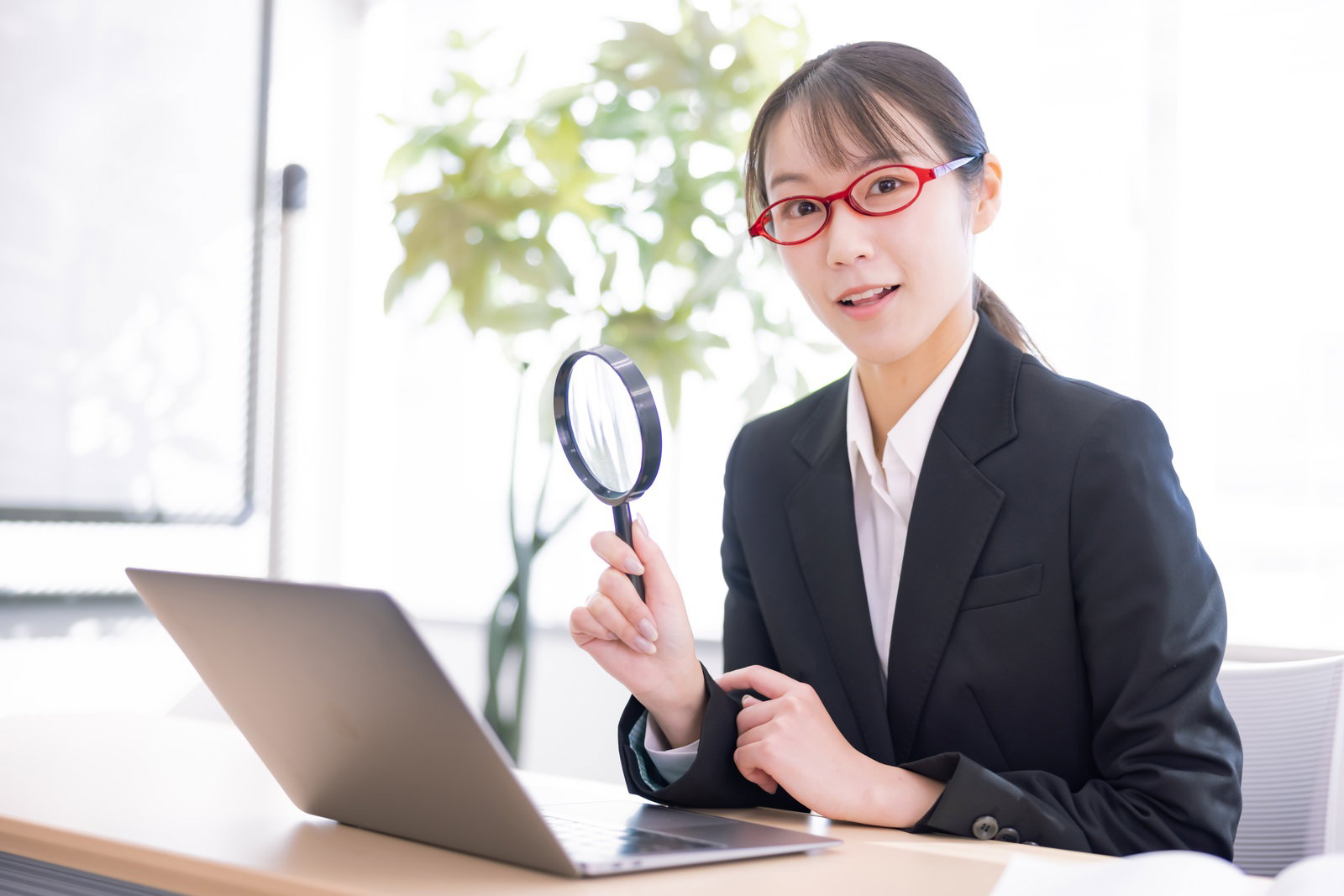 オフィスで虫眼鏡を持ちながら検索作業をする黒いスーツの女性［モデル：SAKI］