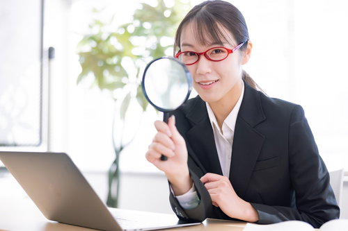 拡大鏡を持ってパソコン画面を検索する、メガネをかけた女性会社員