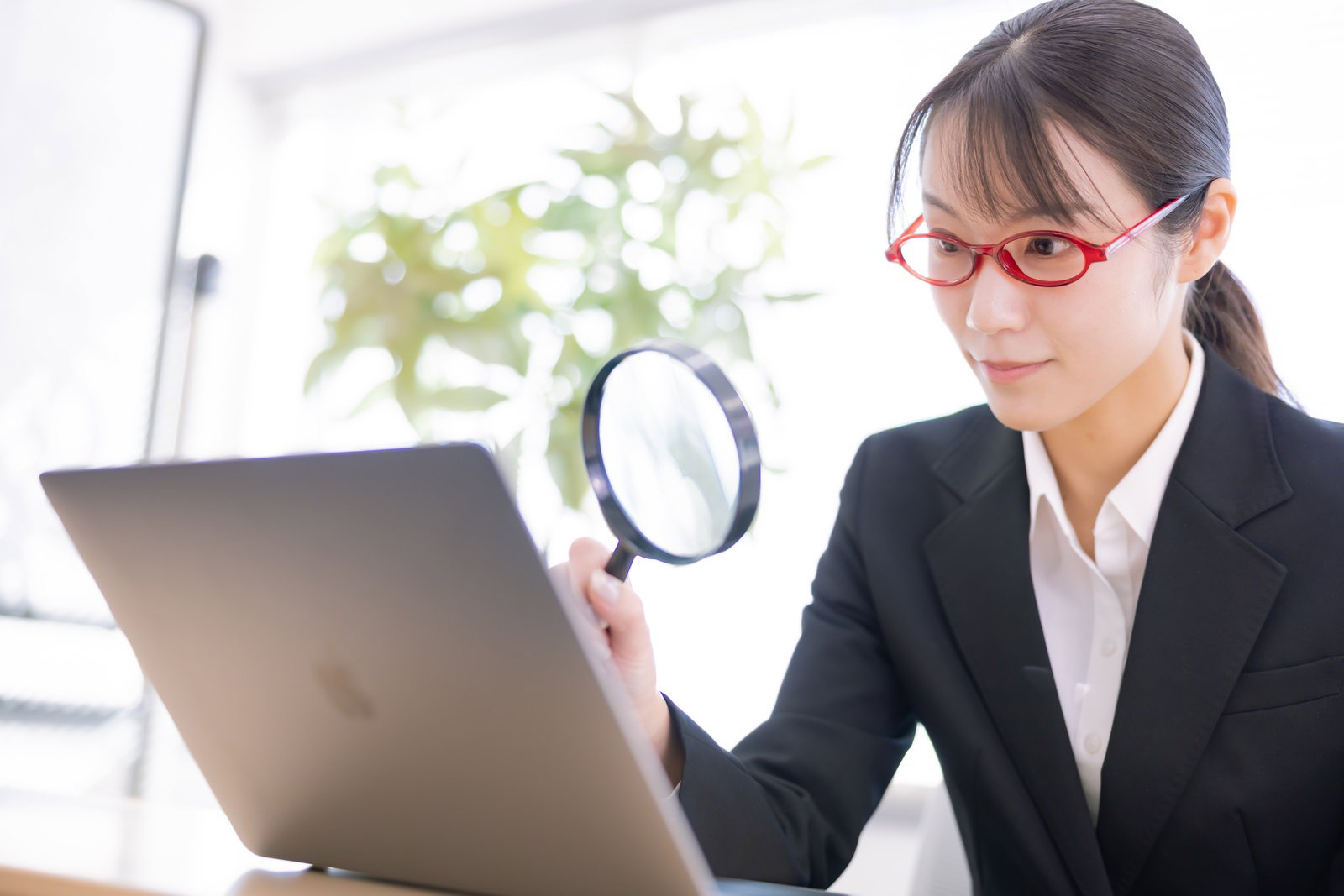 赤い眼鏡の女性社員が虫眼鏡を持ちながらパソコンを操作している［モデル：SAKI］