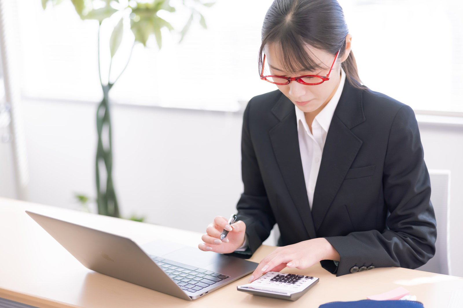 赤いメガネをかけた女性会社員がノートパソコンの横で電卓を操作して計算している［モデル：SAKI］