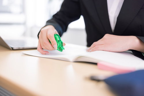 オフィスの机上で書類に修正テープを当てて訂正作業をする女性の手元