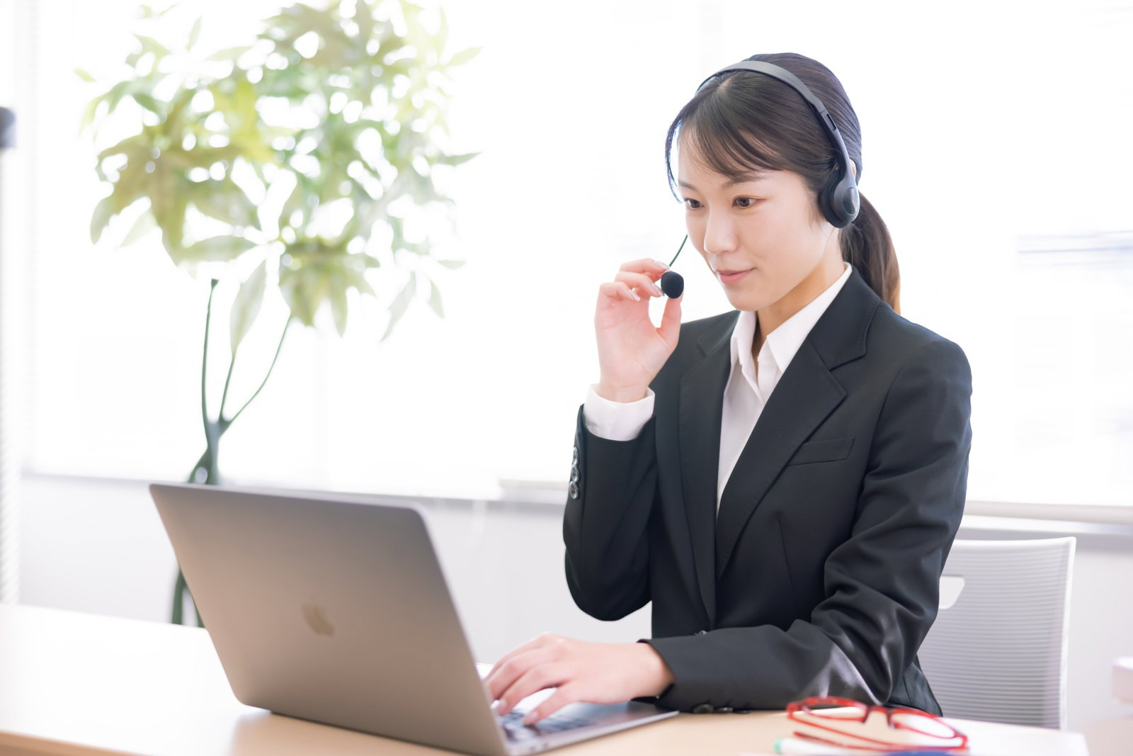 ヘッドセットを装着してノートパソコンで作業する黒いスーツの女性［モデル：SAKI］
