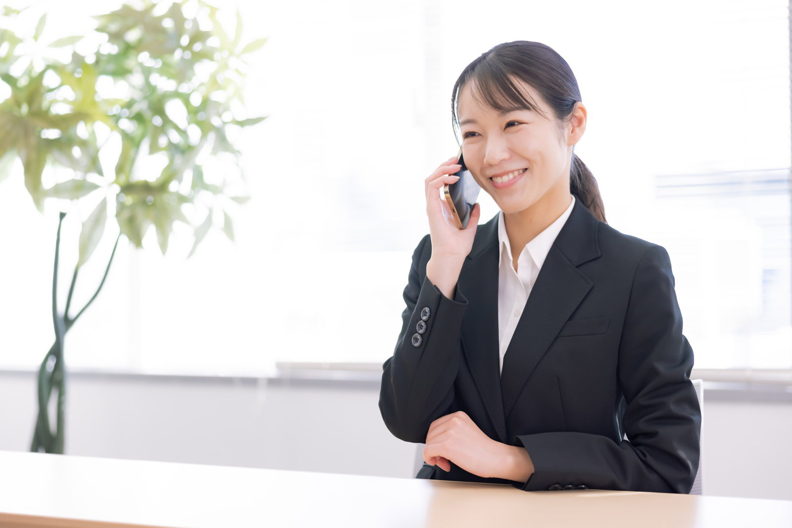 オフィスのテーブルでスマートフォンで通話する笑顔の女性事務員［モデル：SAKI］