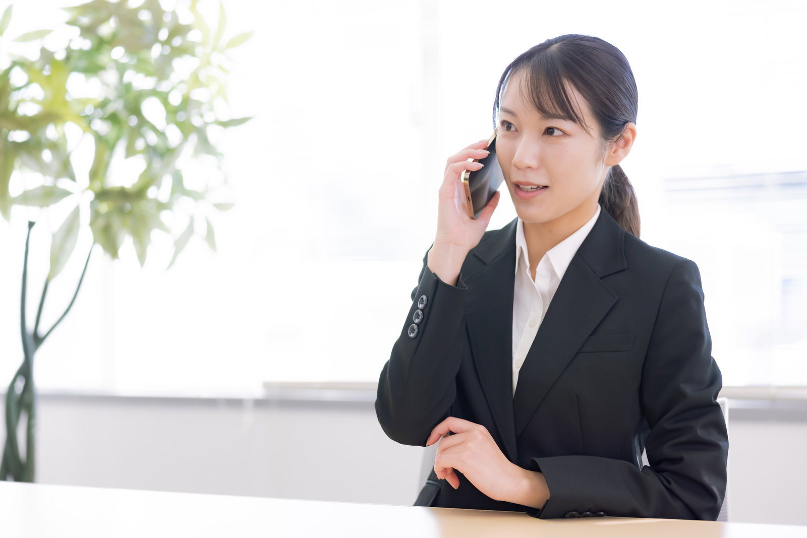 黒いスーツを着た女性事務員がオフィスでスマートフォンで通話している［モデル：SAKI］