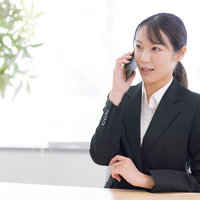 スマホで通話中の女性事務員がデスクで対応する業務風景の写真