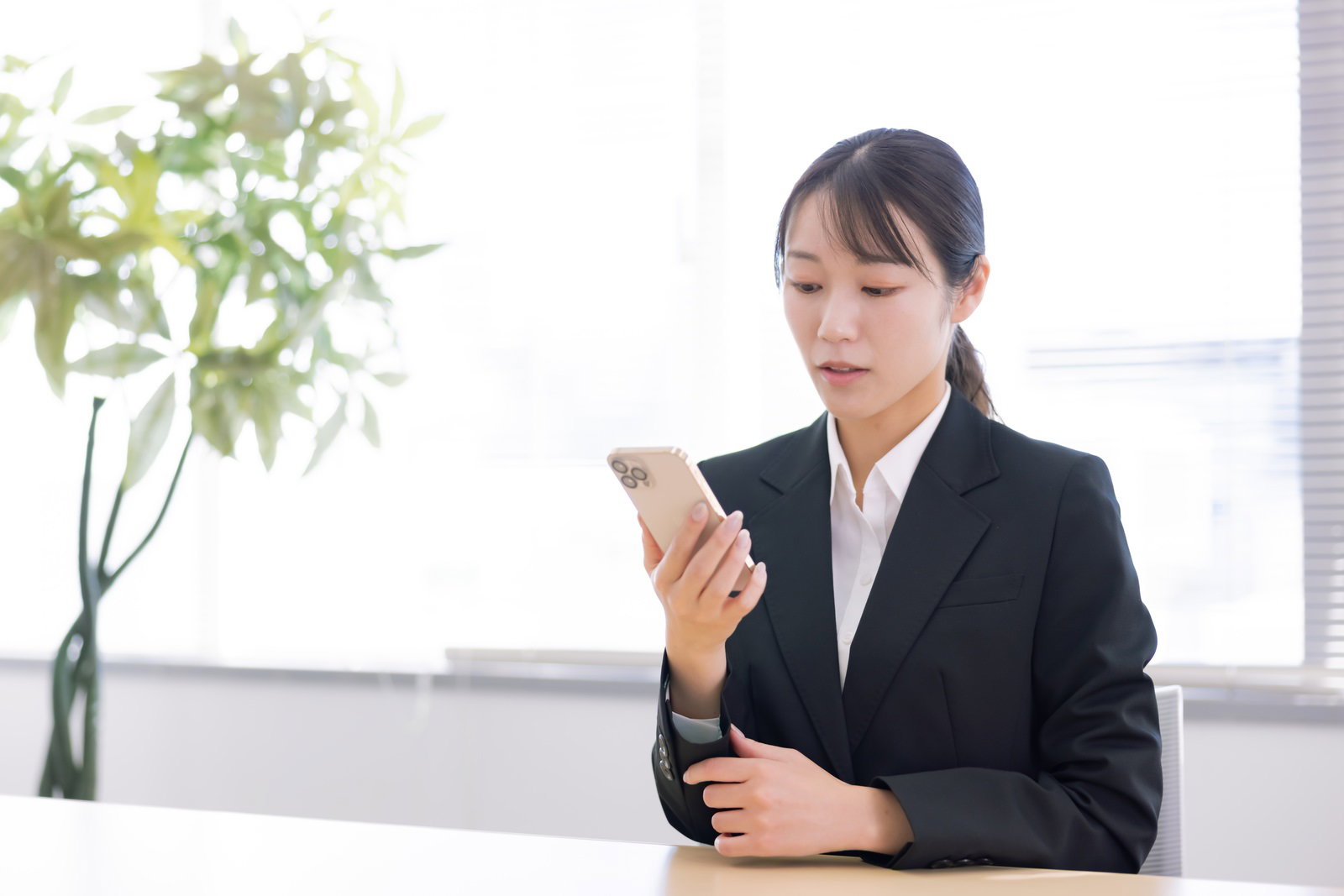 オフィスのデスクでスマートフォンを手に持ち画面を見つめる黒いスーツ姿の女性新入社員［モデル：SAKI］