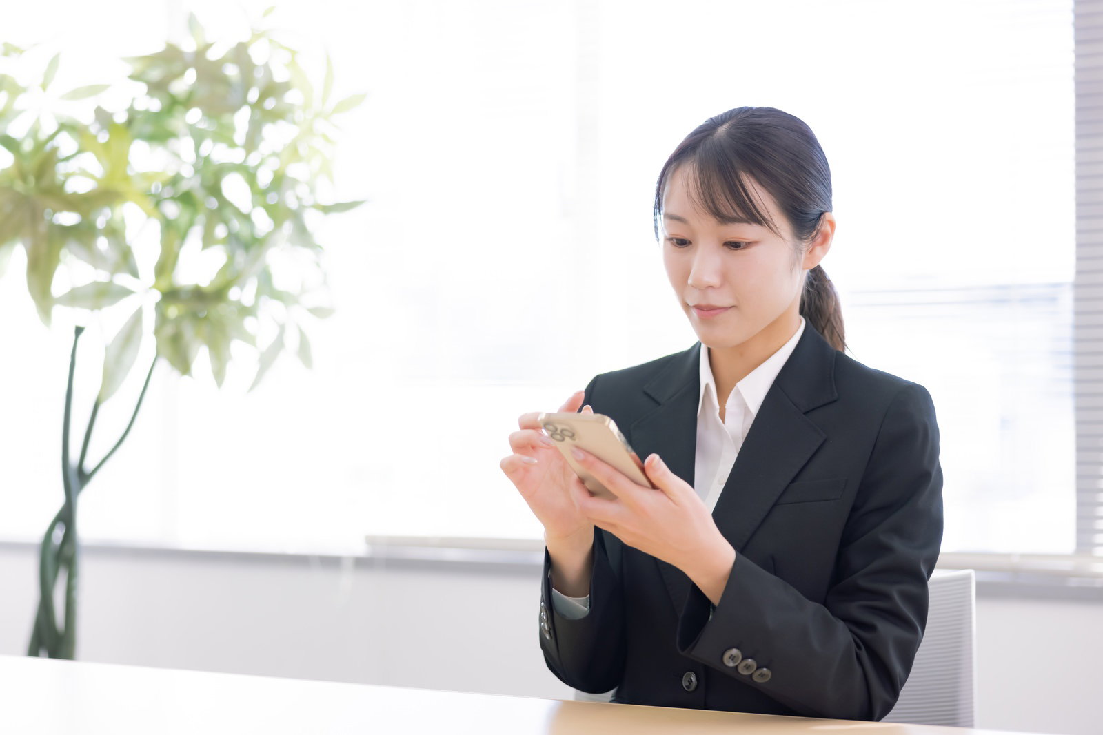 黒いビジネススーツを着た女性がオフィスでスマホを操作している［モデル：SAKI］