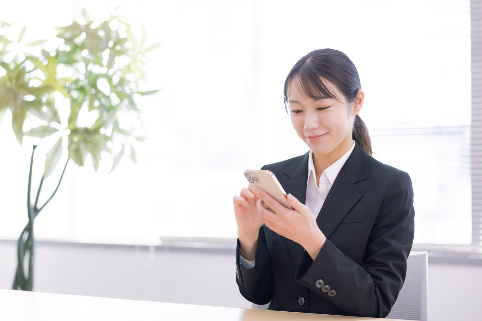 スマートフォンを持ち、画面を見て微笑む黒いスーツ姿の女性新入社員［モデル：SAKI］
