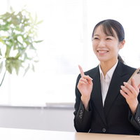 スマホを持ちながら笑顔で人指し指を立てる女性の写真