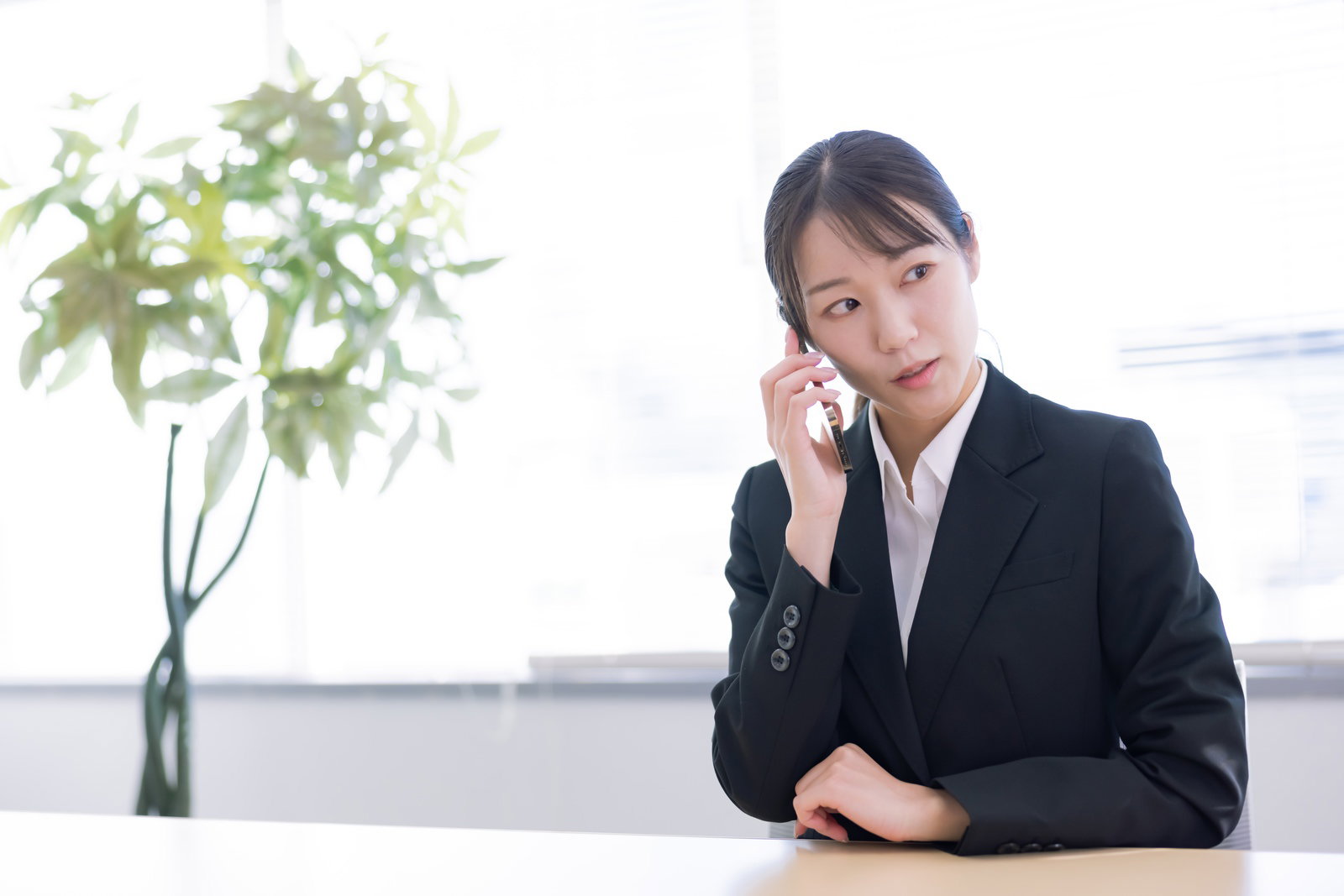 机に座り電話で通話をする黒いスーツ姿の女性［モデル：SAKI］