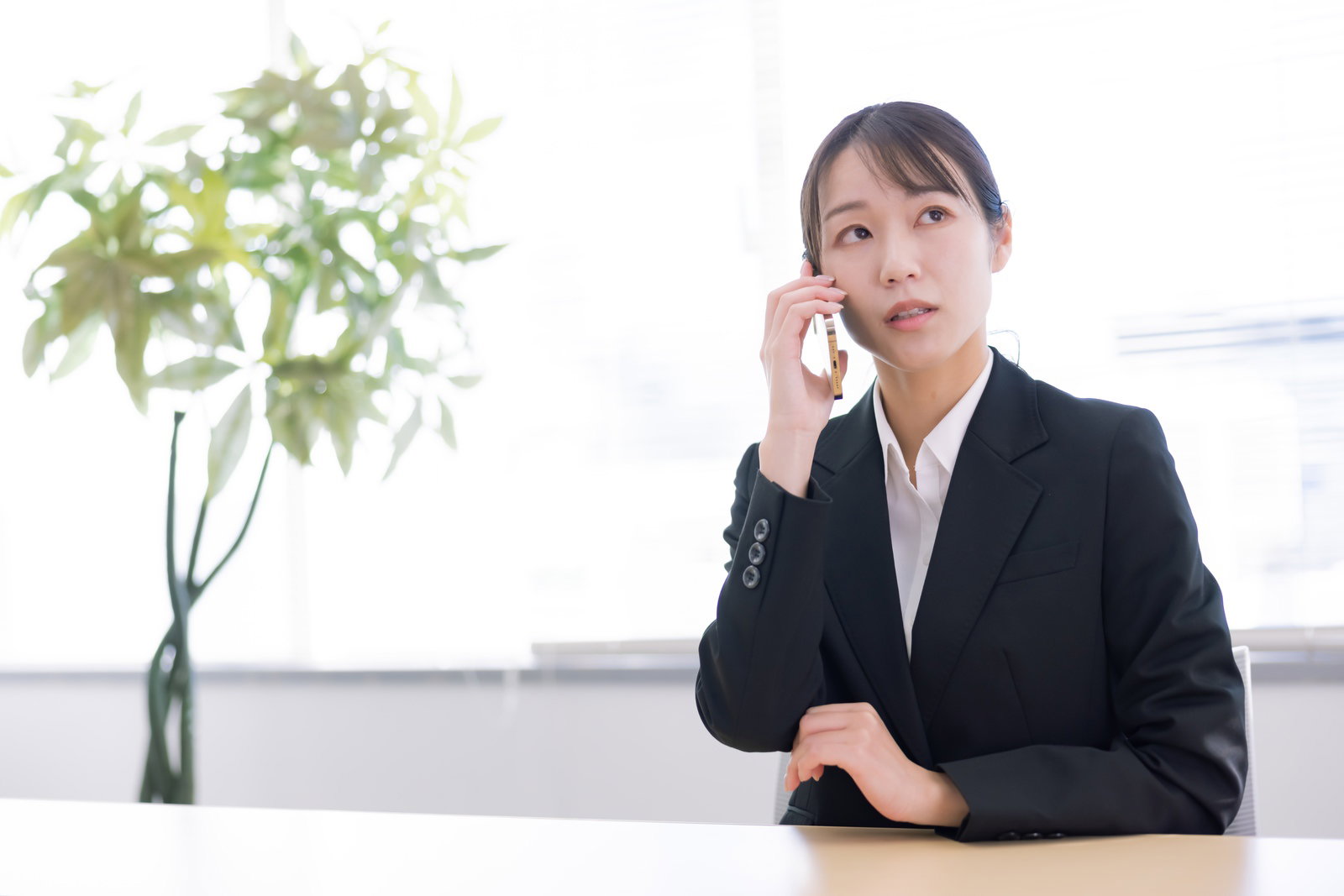 黒いスーツを着た女性が机に座ってスマートフォンで通話している［モデル：SAKI］