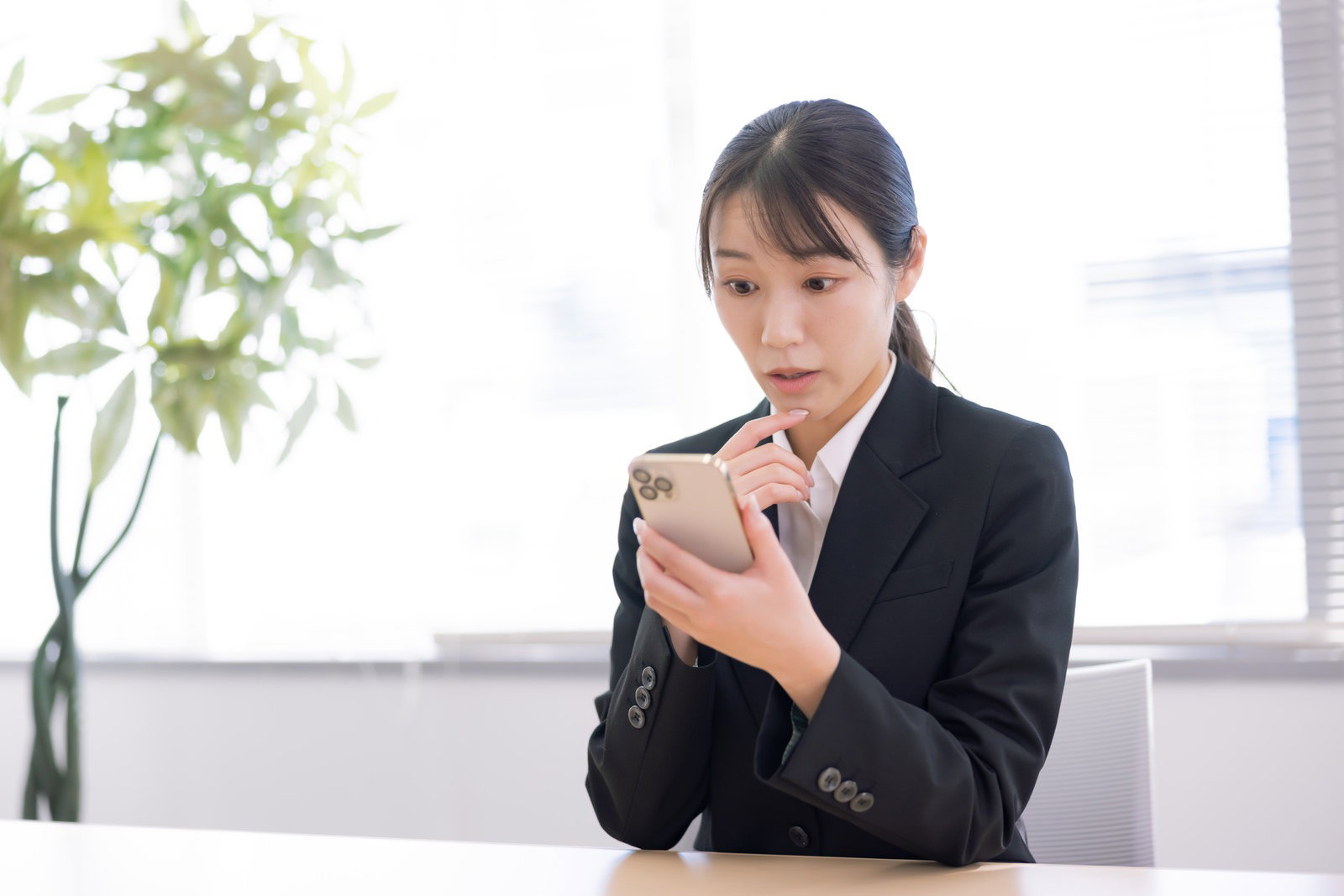 黒スーツの女性事務員がデスクでスマートフォンを見て驚いている様子［モデル：SAKI］