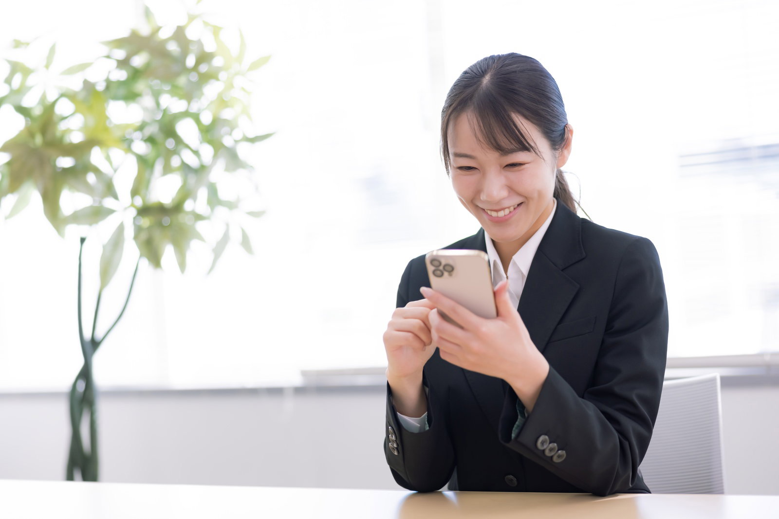 黒いスーツを着た女性がオフィスのデスクでスマートフォンを見て笑顔を浮かべている［モデル：SAKI］
