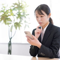 スマホを見て首をかしげ考え込む女性事務員の写真
