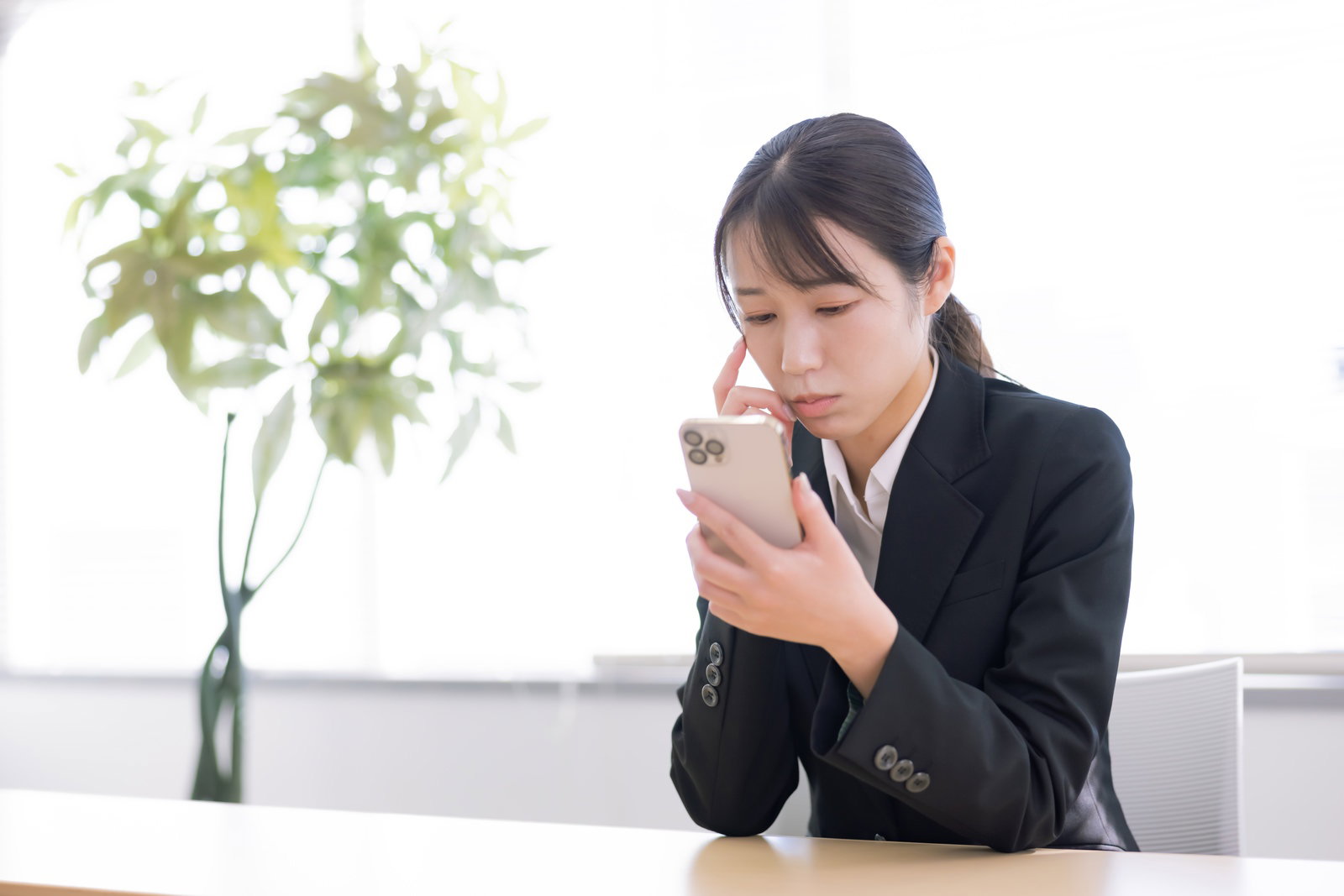 A woman in a black suit is holding a smartphone and has her finger on her chin, looking at the screen with a confused expression.