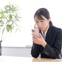 スマートフォンの操作に戸惑う黒いスーツの女性事務員の写真