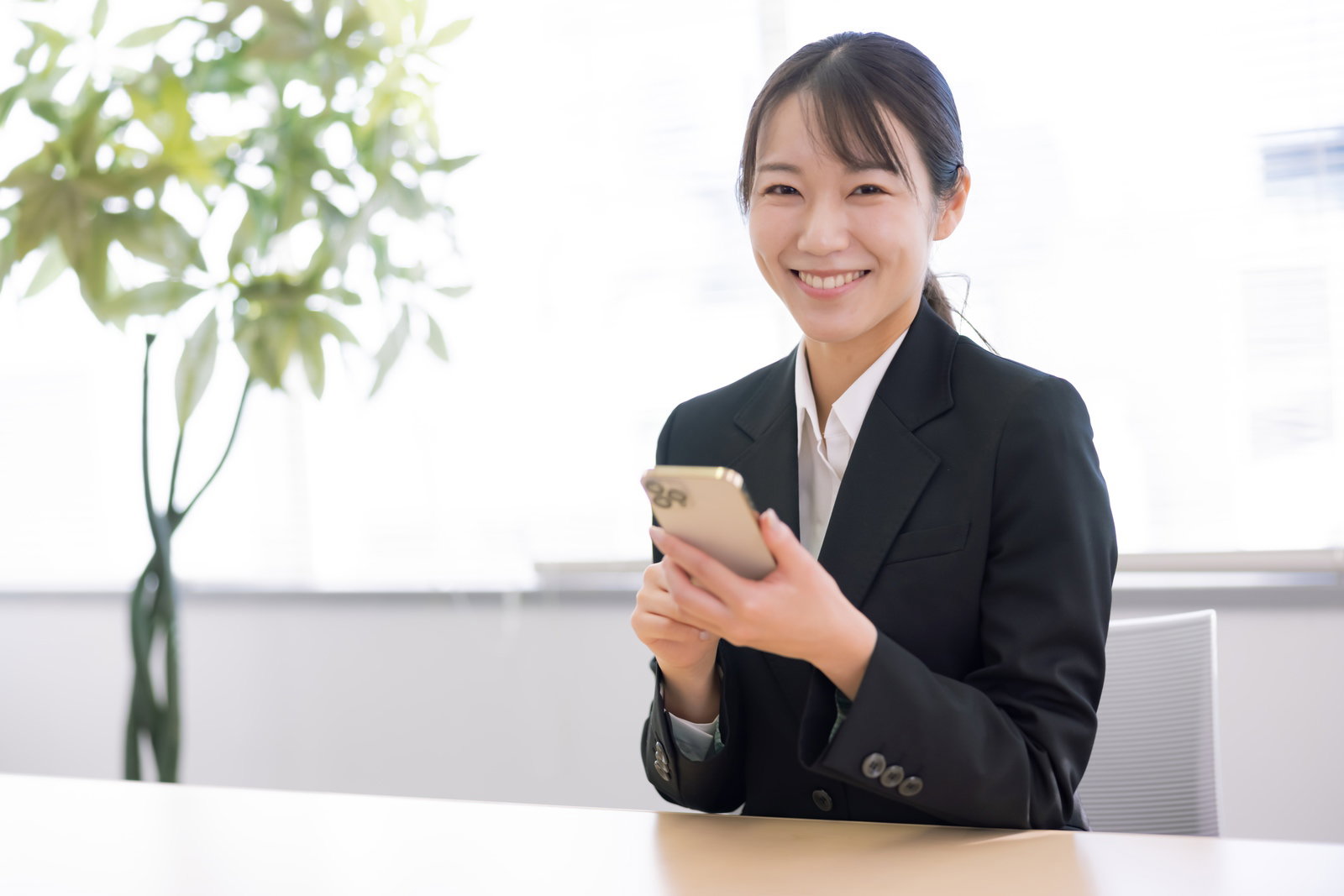 A woman in a black suit is smiling and holding a smartphone at an office desk