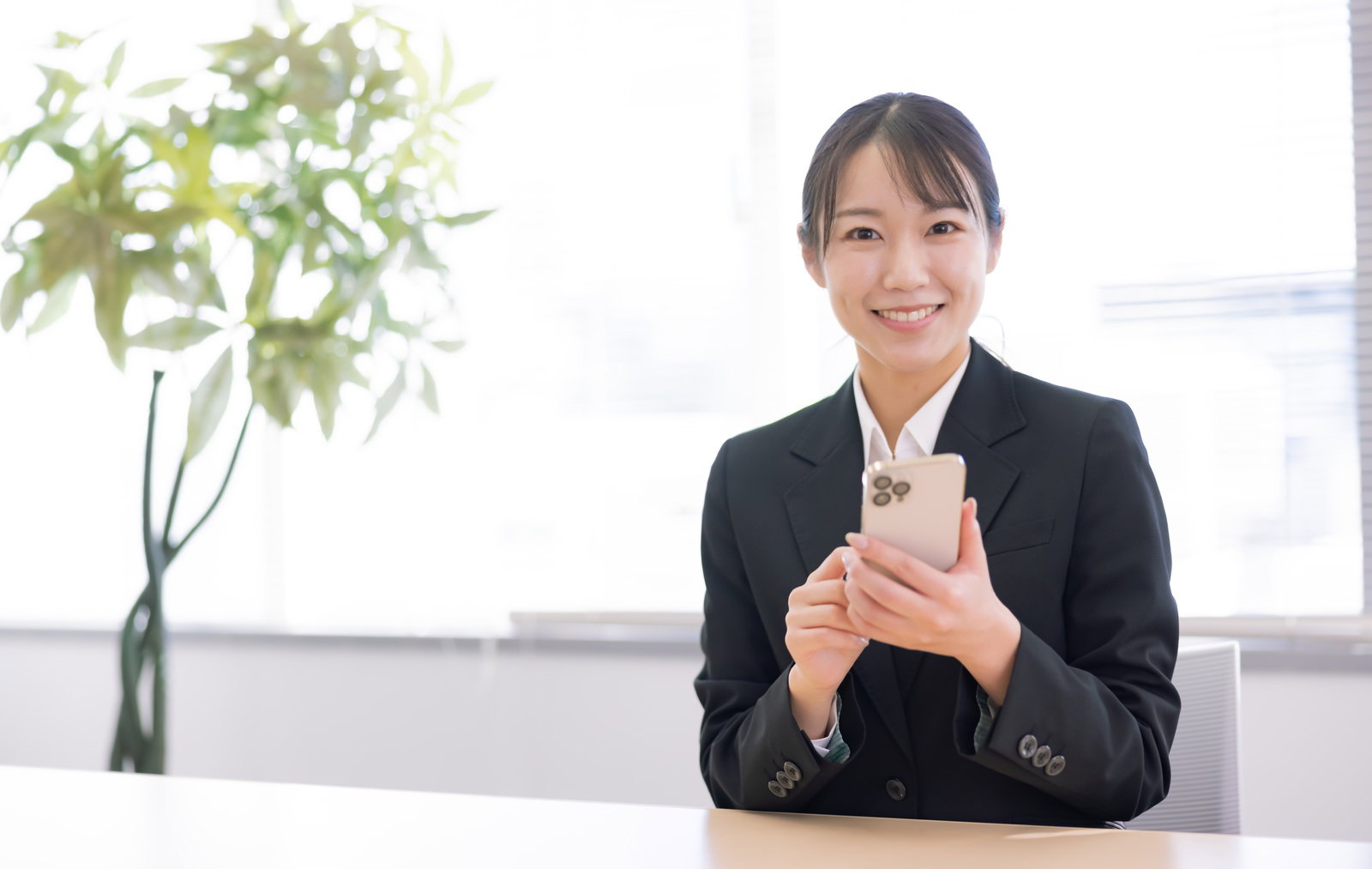 オフィスのテーブルでスマートフォンを持ち笑顔を見せる黒いスーツの女性会社員［モデル：SAKI］