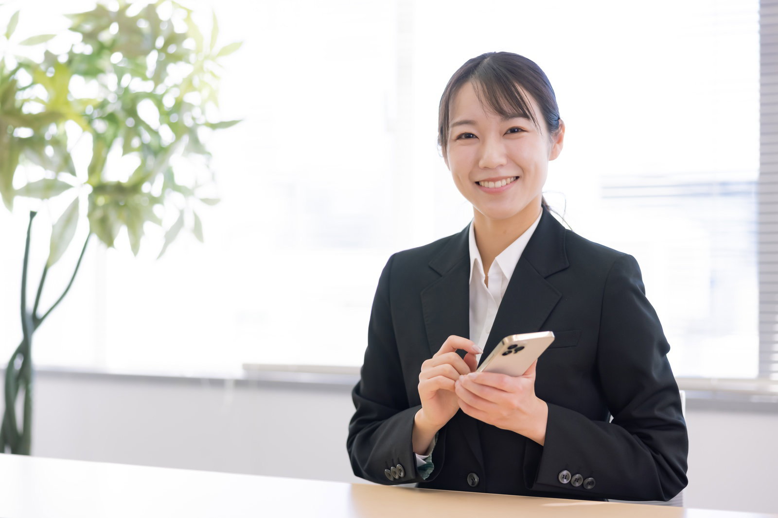 黒いスーツを着た女性店員が笑顔でスマートフォンを操作している［モデル：SAKI］