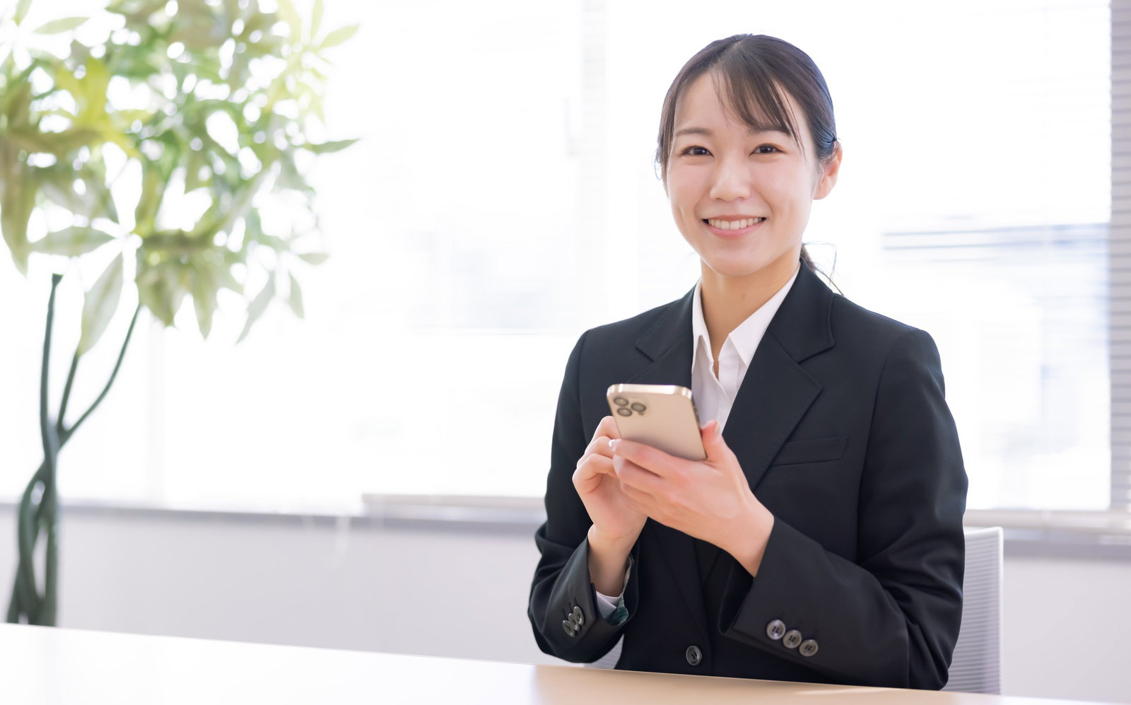 黒いスーツを着てスマートフォンを持ちながら笑顔を見せる女性社員［モデル：SAKI］