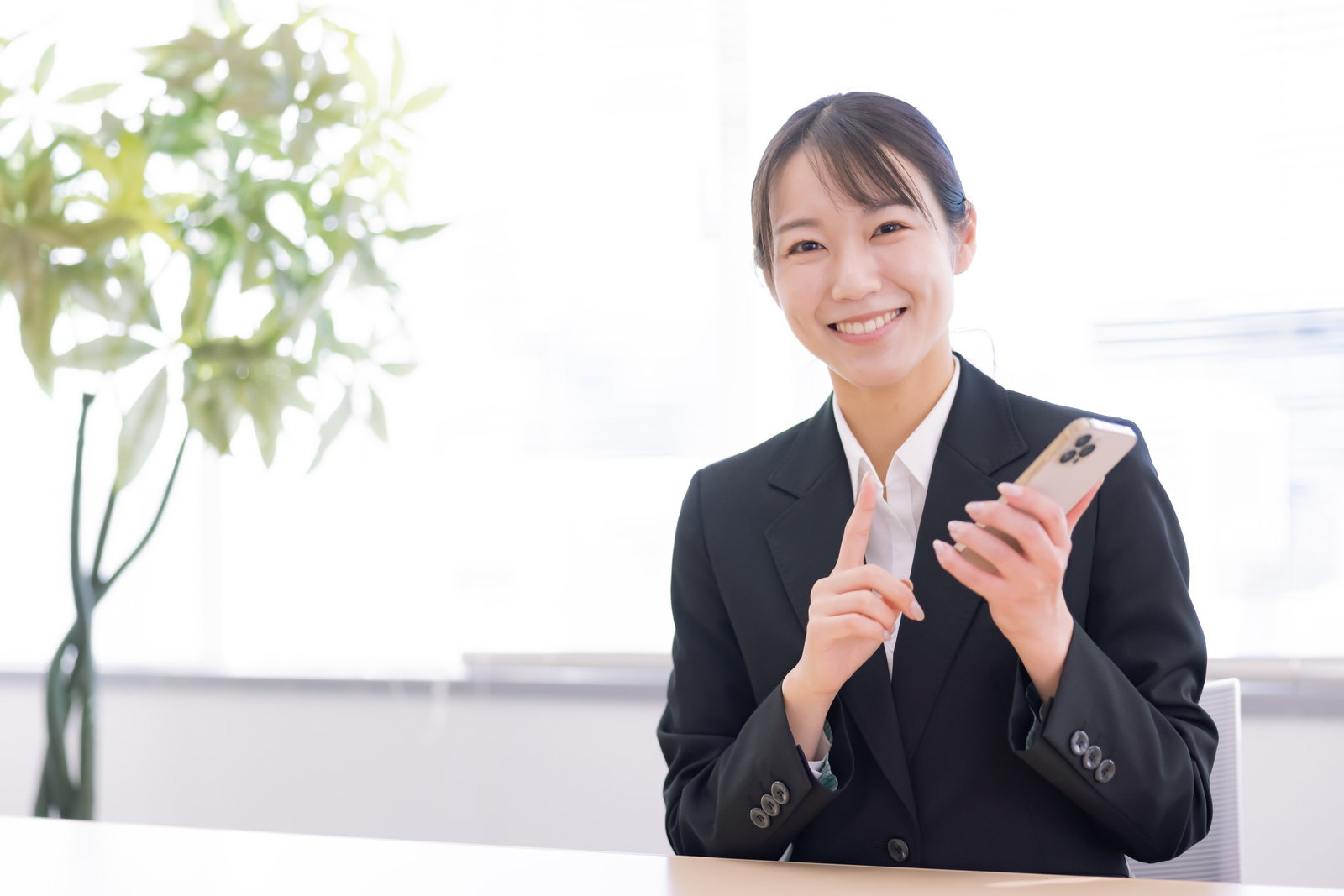 黒いスーツを着た女性がスマートフォンを持って笑顔で説明している［モデル：SAKI］