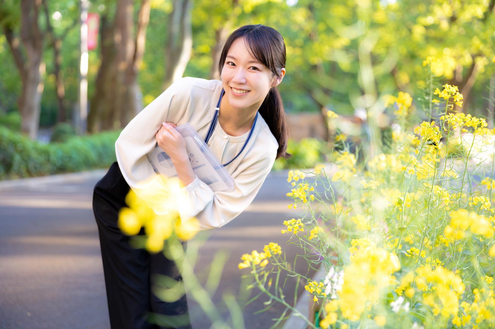 菜の花畑で身を乗り出して手を振るベージュジャケットの女性［モデル：SAKI］