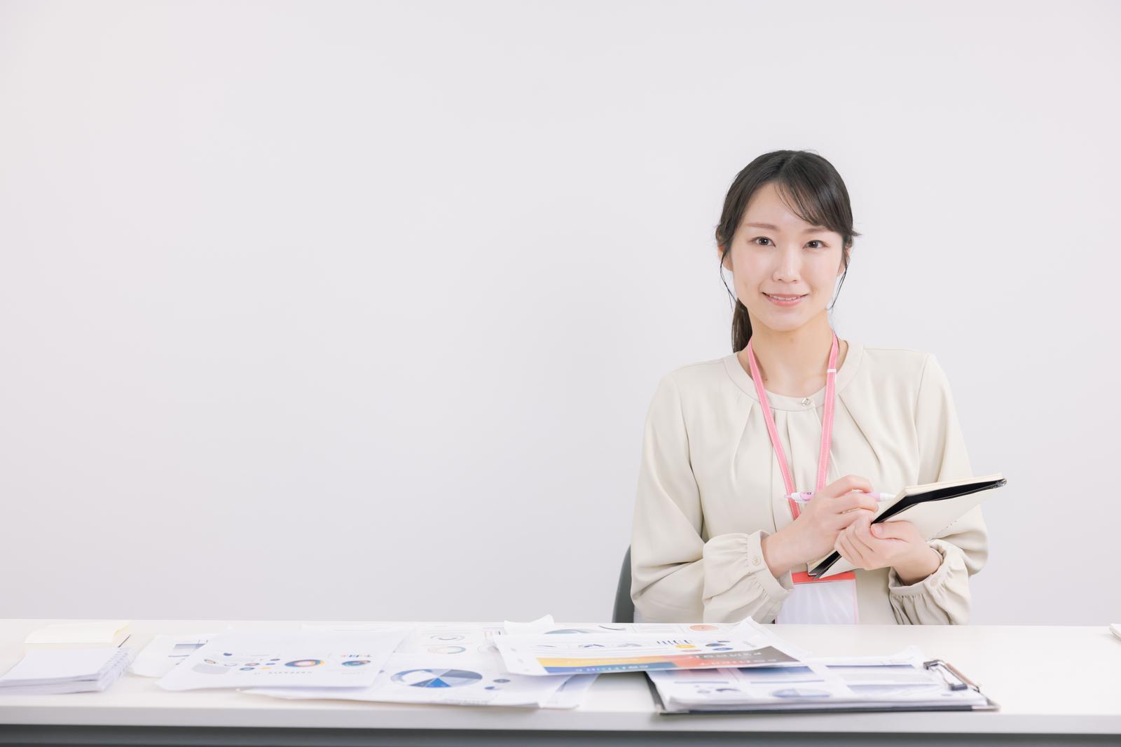 デスク上の書類を確認しながら議事録をとる事務職の女性［モデル：SAKI］
