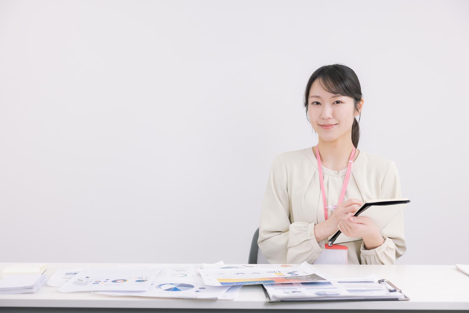 タブレットとメモ帳を持つビジネスウーマンが書類に囲まれて作業中［モデル：SAKI］