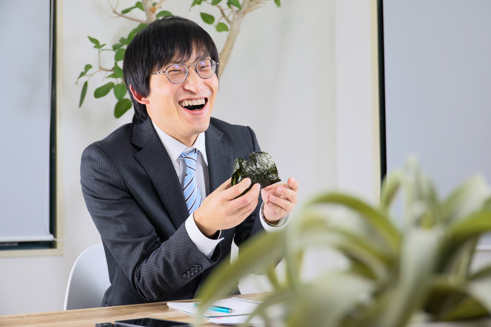 会議室でおにぎりを食べながら笑う眼鏡の男性社員［モデル：あづま］