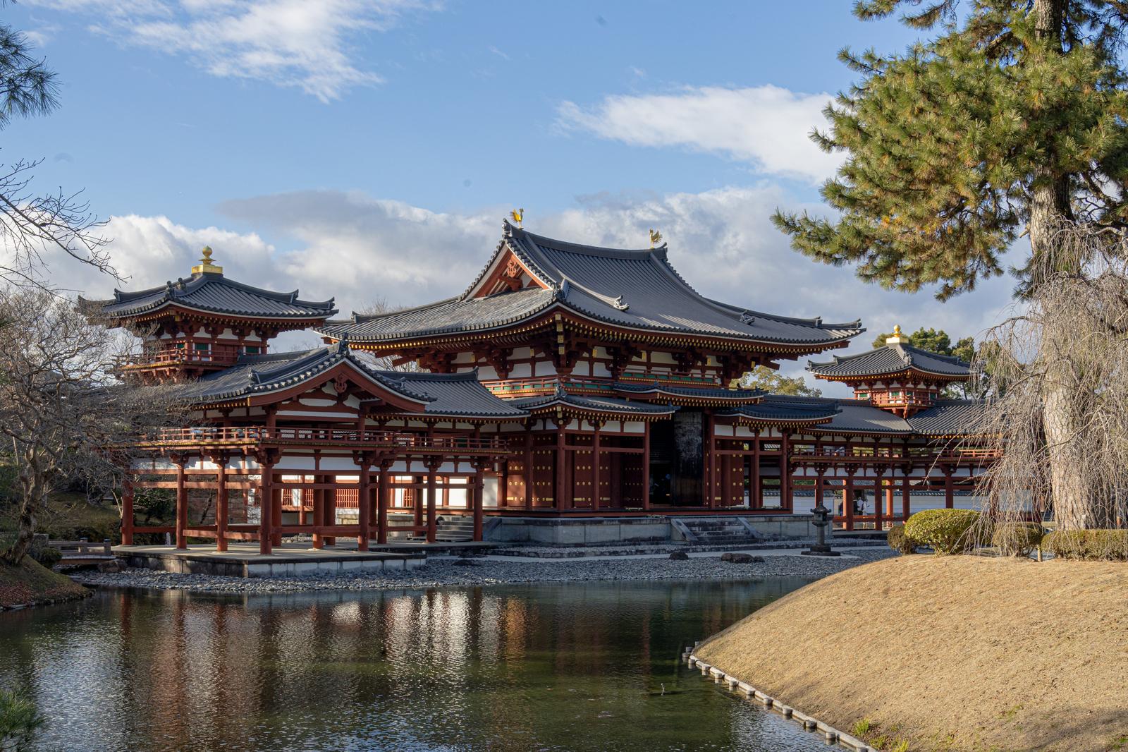 Byodoin Phoenix Hall in Uji, Kyoto: vermilion columns, grey tiled curved roofs, and gilded ornaments reflected in a surrounding pond under blue sky.