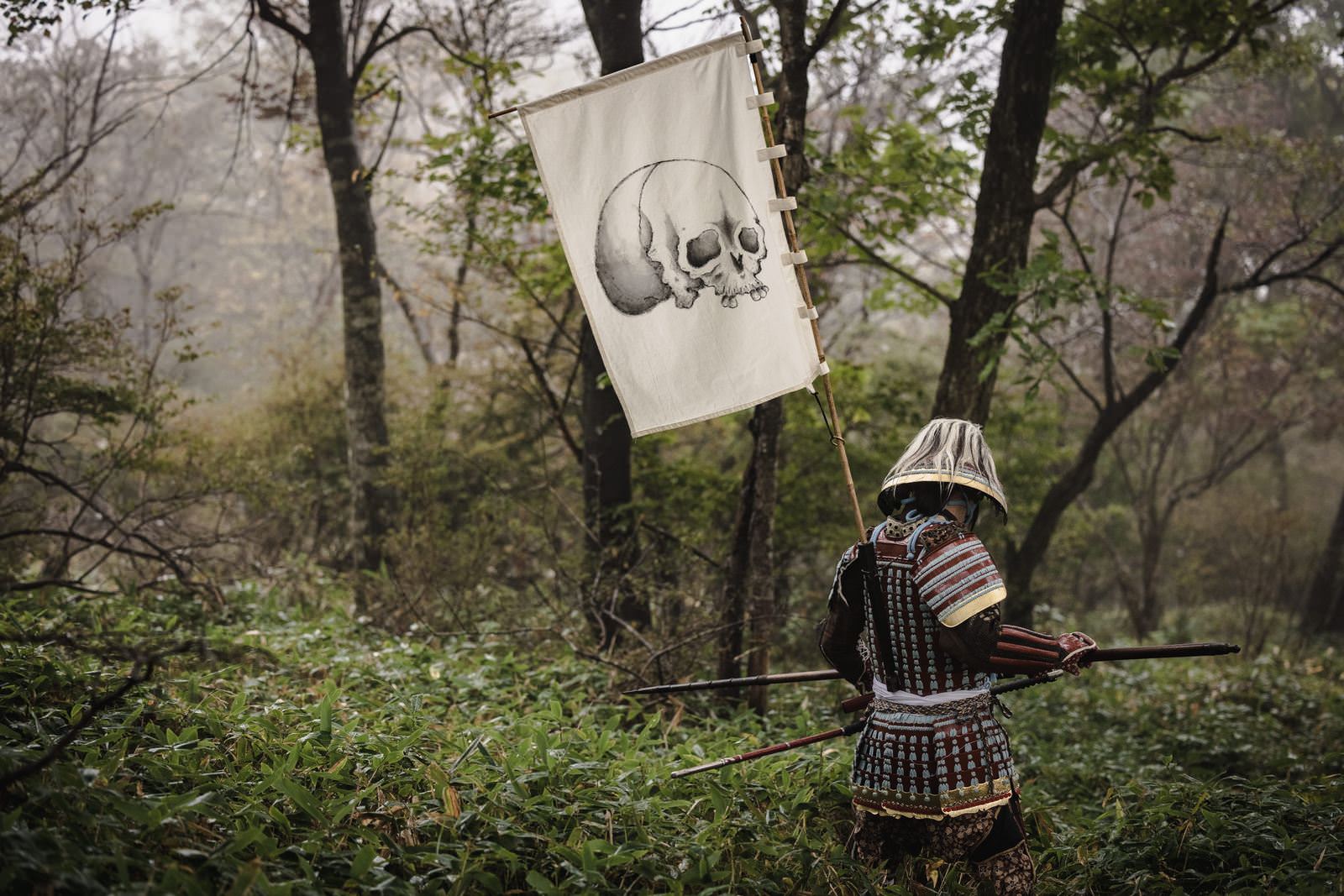 骸骨の指物を背負い、黒い甲冑を着た武士が森の中に立っている［モデル：まーこ］