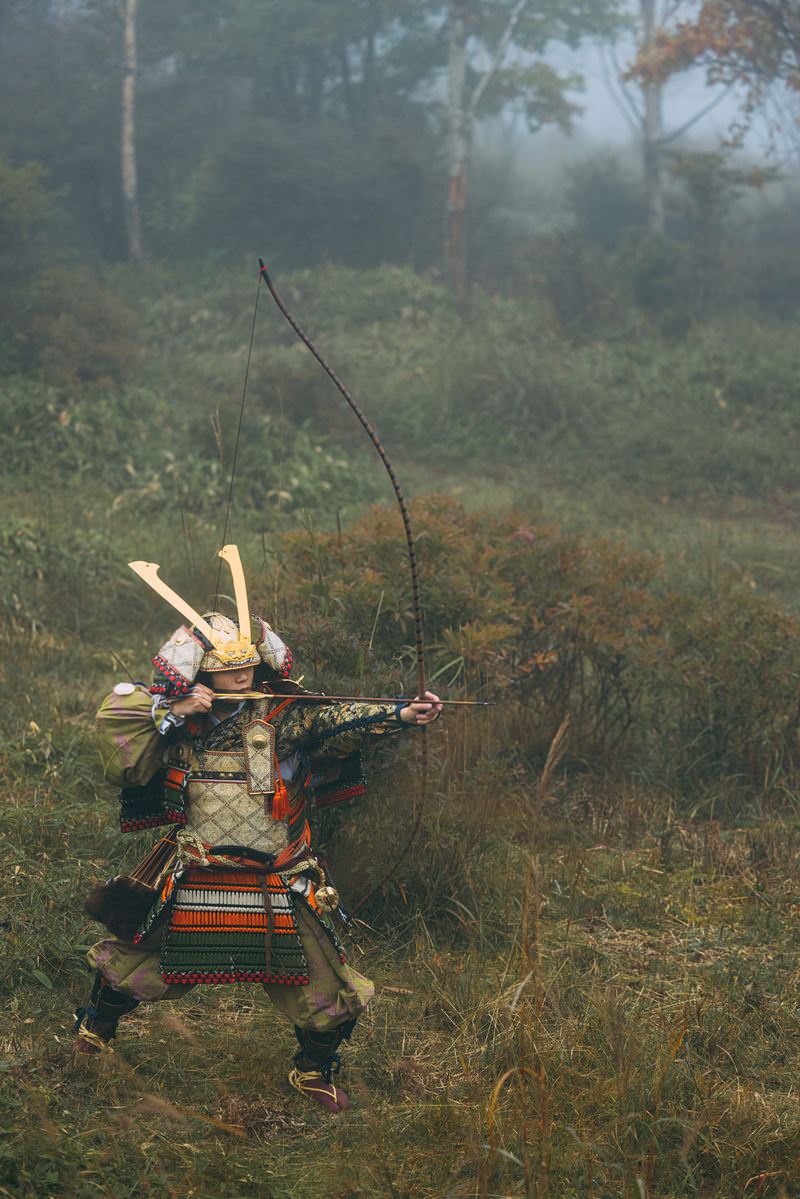 霧の立ち込める野原で大弓を引く鎧姿の侍［モデル：まーこ］