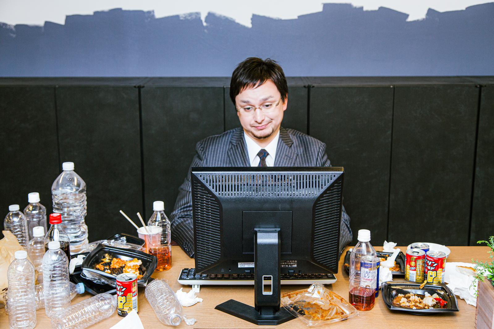 徹夜残業でデスクが散らかるスーツ姿の男性とコンビニ弁当［モデル：Max_Ezaki］