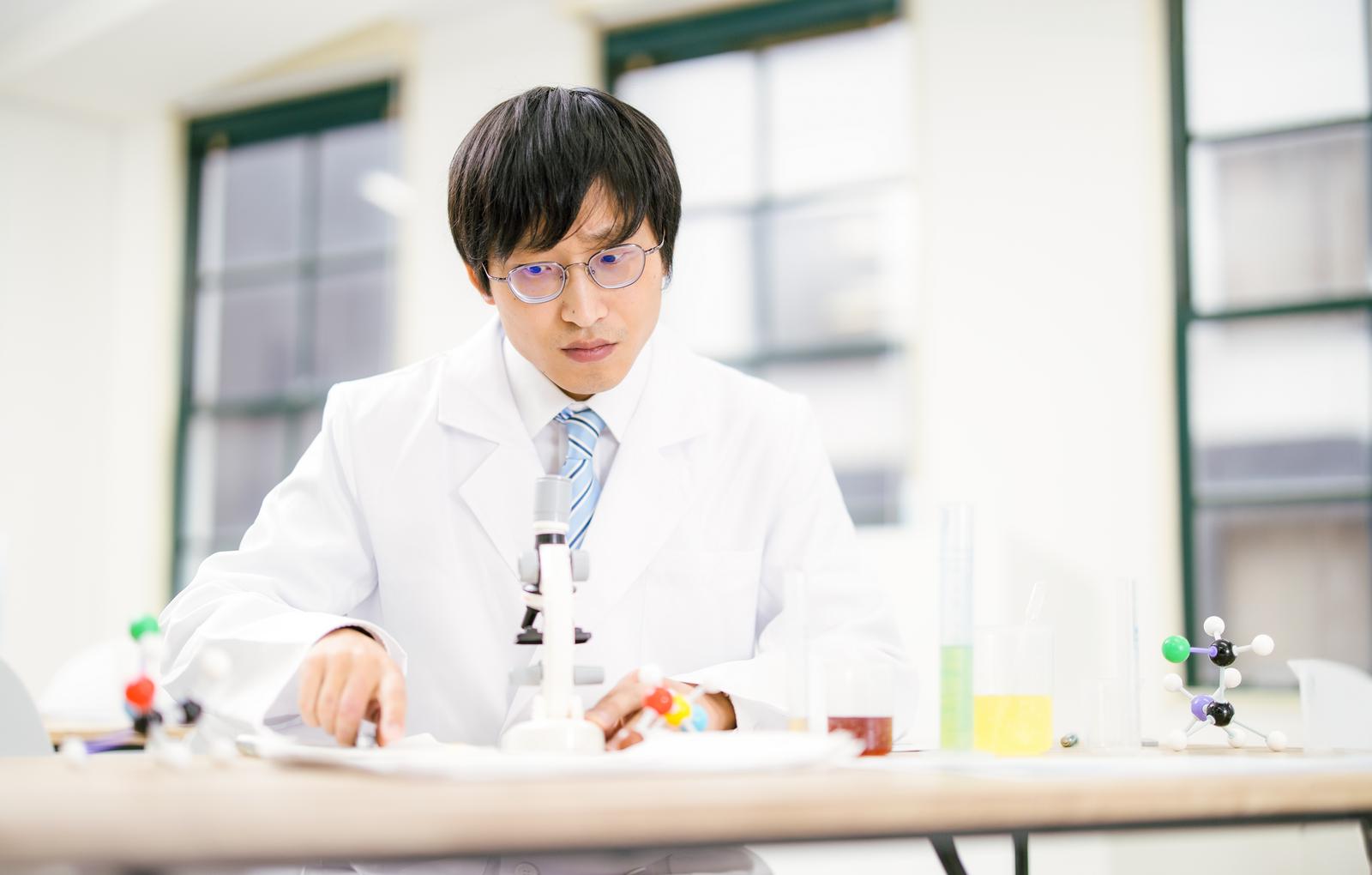 白衣を着た眼鏡の男性が分子構造模型と顕微鏡を使い実験する研究室での一場面［モデル：あづま］