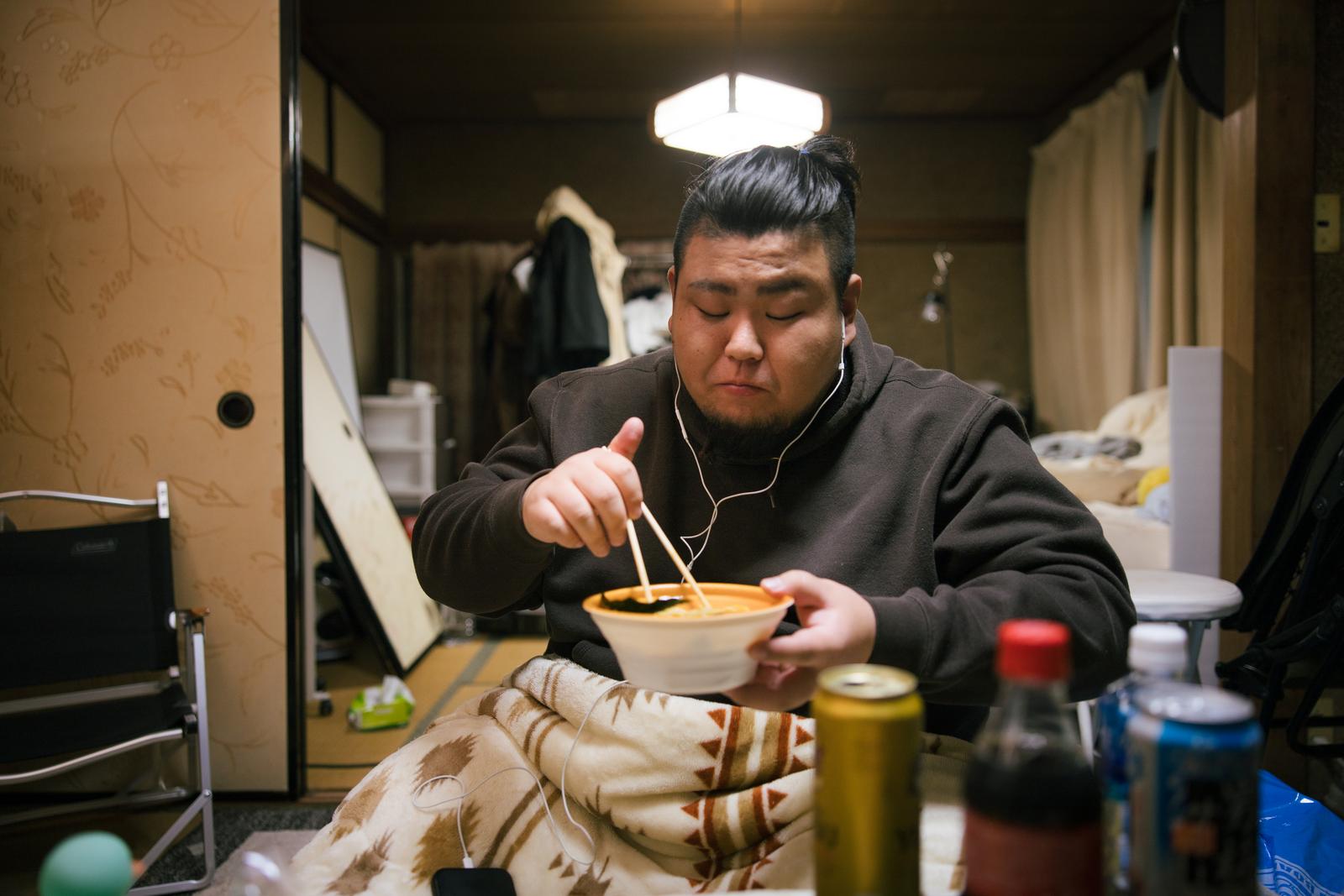 ゴミ屋敷でラーメンを食べる男性がヘッドフォンをして箸で麺をすくっている［モデル：ノリックス］