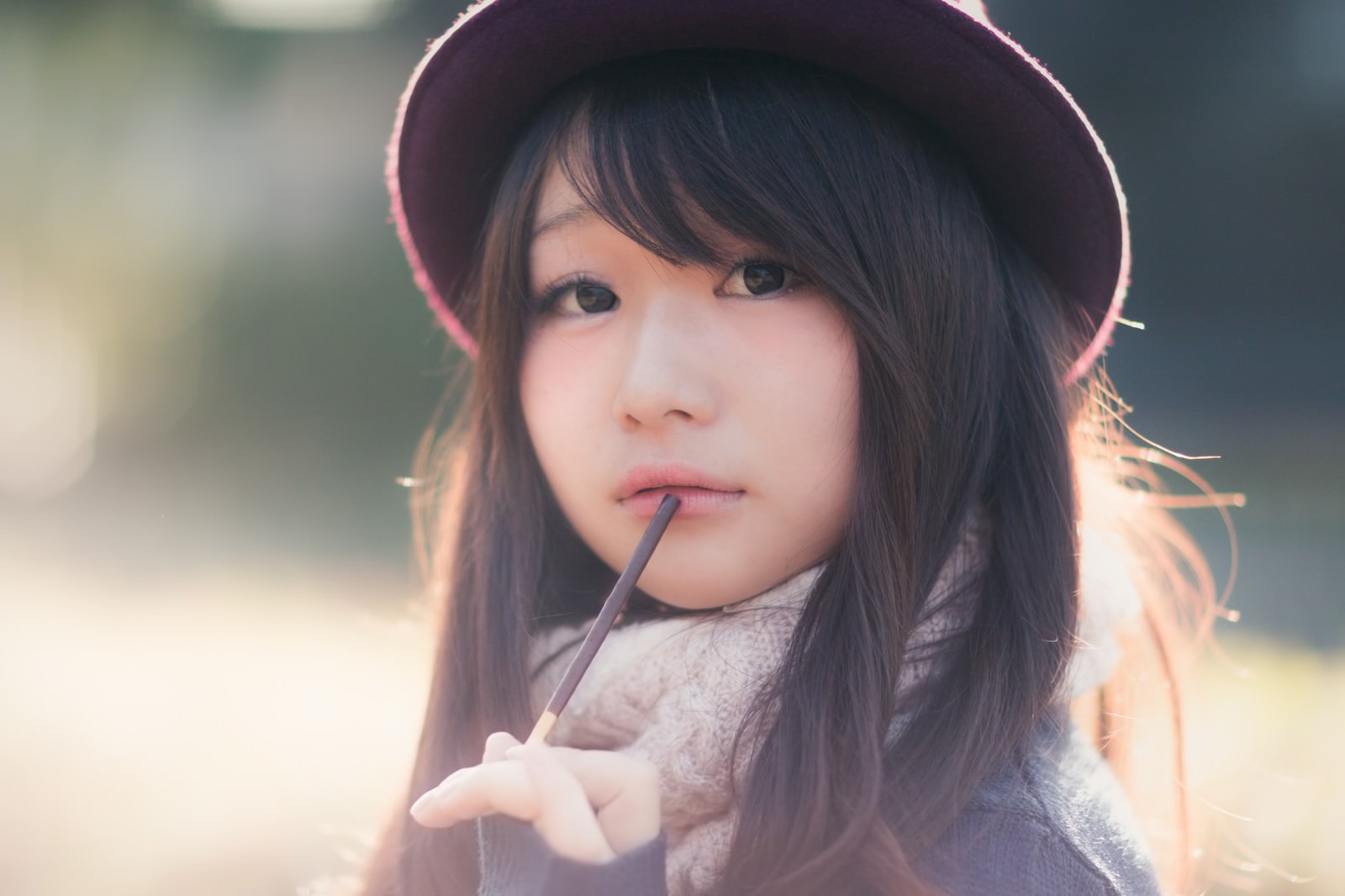 紫色の帽子をかぶり、グレーのマフラーを巻いた女性がチョコレートポッキーを口にくわえ、斜め上を見つめている［モデル：茜さや］