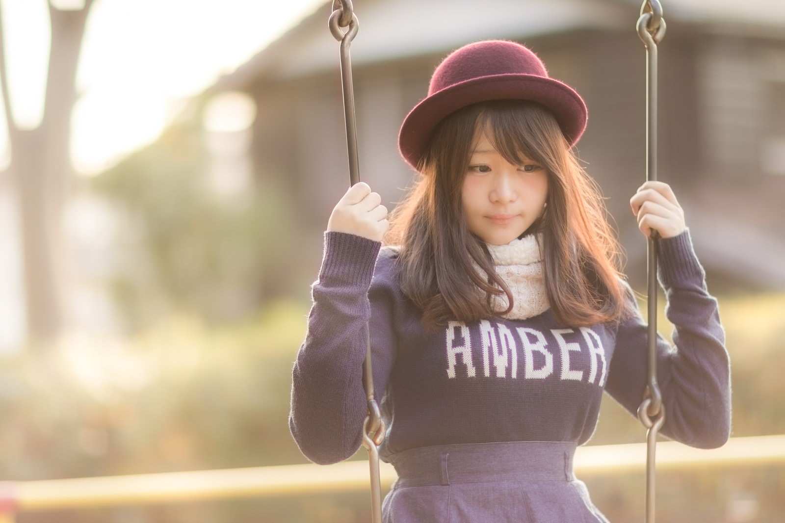 ブランコに乗ってうつむく女性。ピンク帽子とスウェット姿で、夕暮れの光に包まれている［モデル：茜さや］