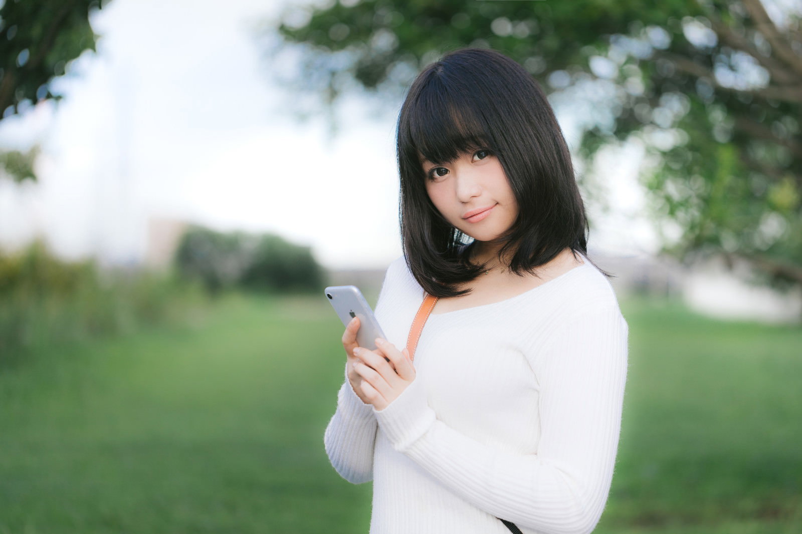 公園でスマートフォンを見つめる白いニットトップスの女性、時間確認の場面［モデル：茜さや］