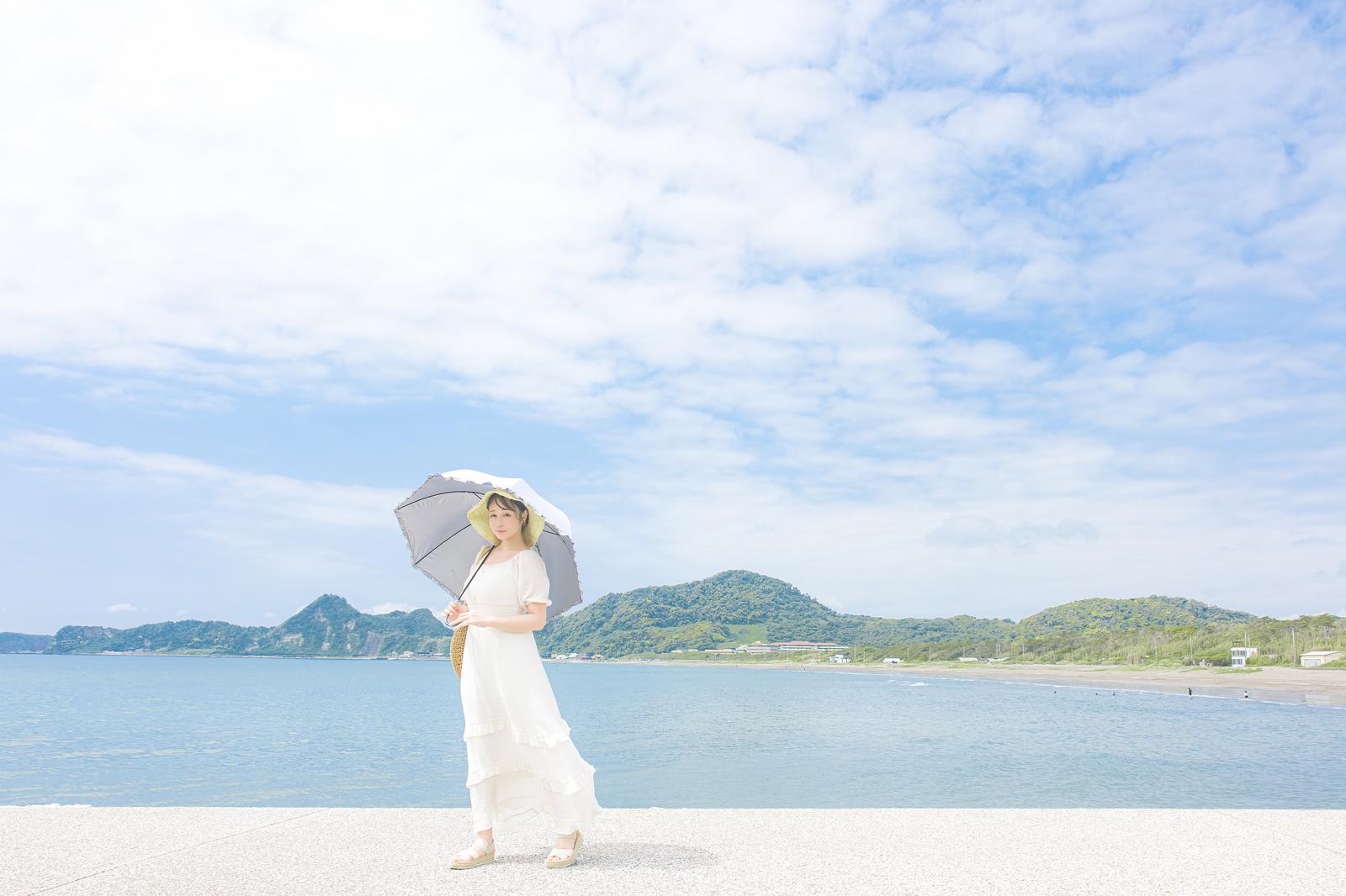 白いワンピースと黄色い帽子で日傘を差す女性が、青い海を前に防波堤に立っている［モデル：茜さや］