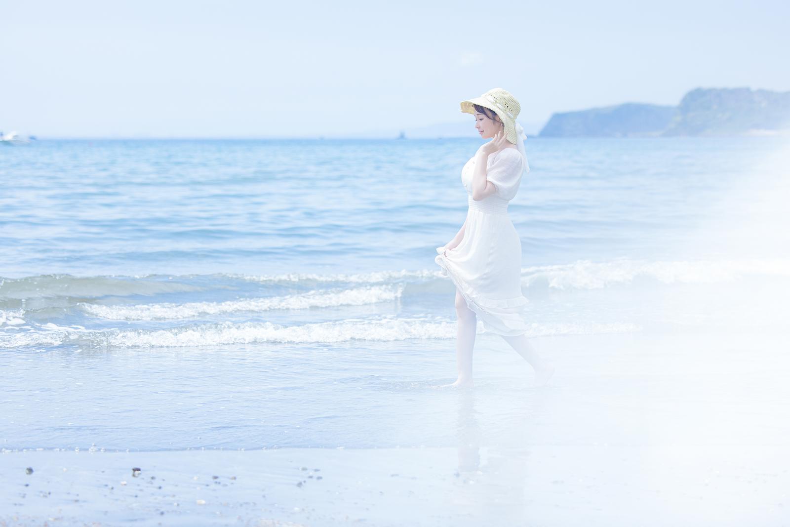 白いワンピースと麦わら帽子を着た女性が波打ち際に立っている［モデル：茜さや］