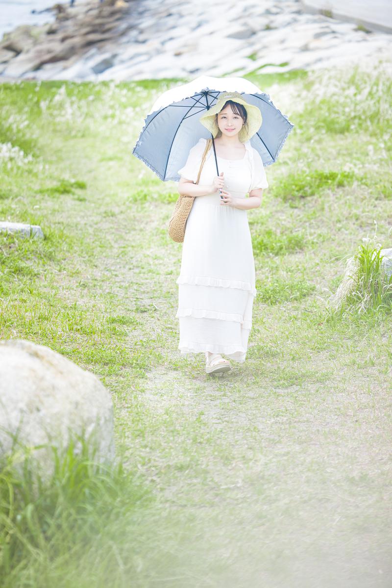 白いワンピースを着た女性が紺色の日傘をさして海岸の草地を散歩している［モデル：茜さや］