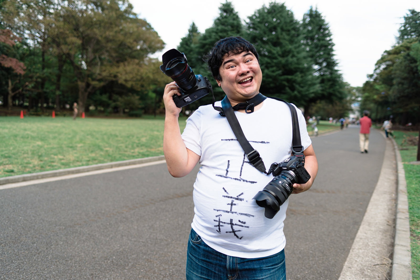 ミラーレスカメラを構えて笑顔で撮影準備をする白いTシャツの男性カメラマン［モデル：段田隼人］