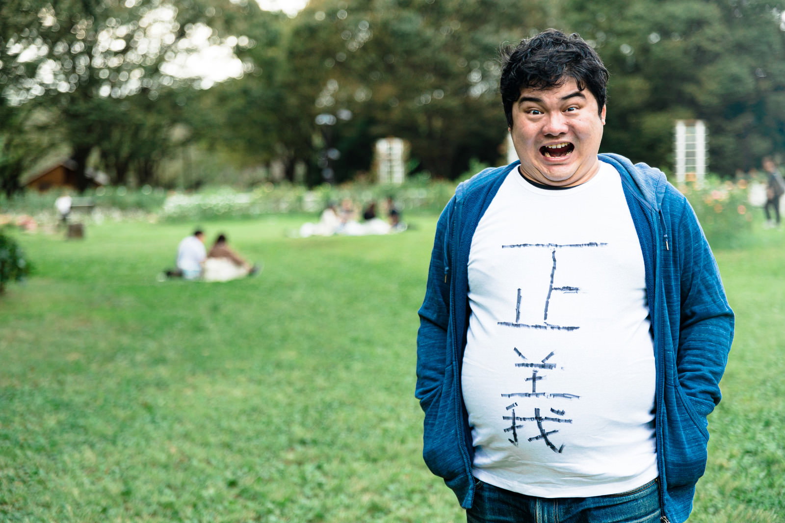 公園の芝生の上で青いカーディガンを着て笑顔を見せる男性［モデル：段田隼人］