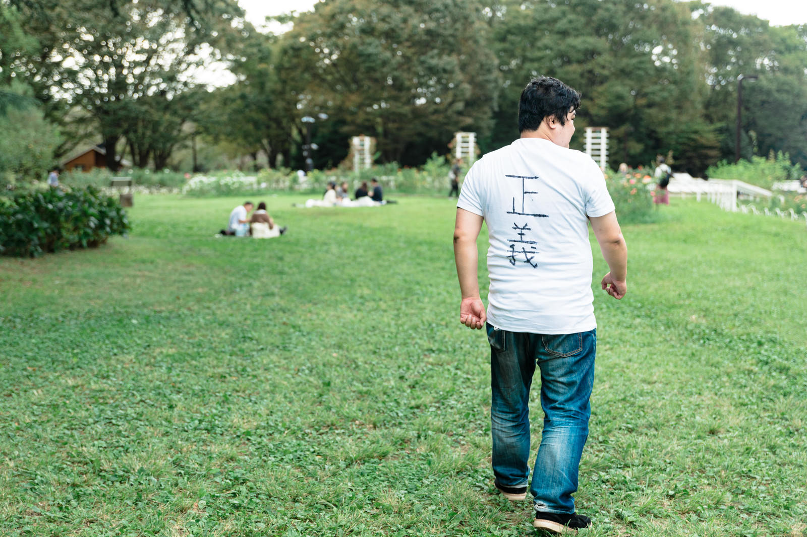 公園の芝生を歩く白いTシャツとジーンズの男性の後ろ姿［モデル：段田隼人］