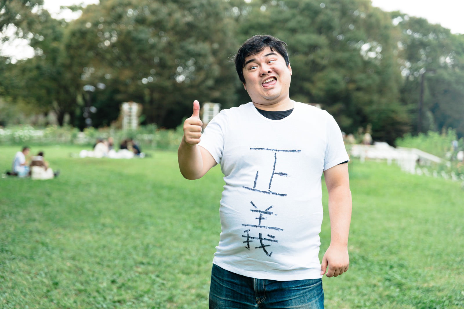 公園の芝生で親指を立ててグッドサインをする白いTシャツの男性［モデル：段田隼人］