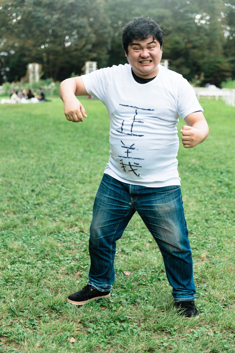 公園の芝生で両手を腰に当ててポーズを決める白Tシャツの男性［モデル：段田隼人］