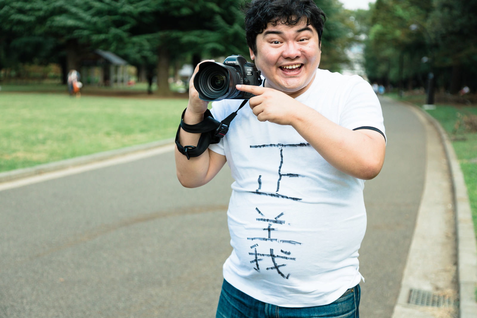 公園で一眼レフカメラを持ちながら笑顔を見せる白シャツの男性［モデル：段田隼人］