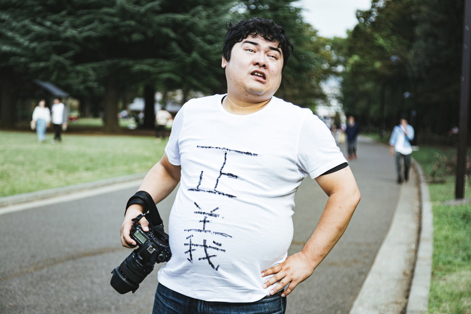 公園の散歩道でカメラを持って立つ白いTシャツの男性［モデル：段田隼人］