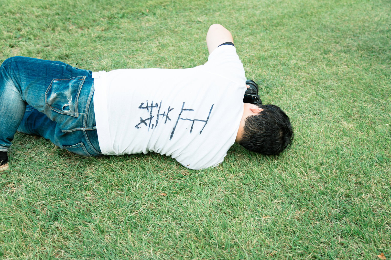 緑の芝生の上に倒れ込んで横たわる、背中に正義マンと書かれた白いTシャツの男性［モデル：段田隼人］